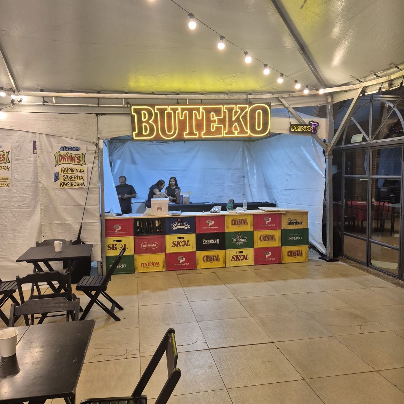Balcão do Buteko com letreiro luminoso e engradados coloridos sob a tenda