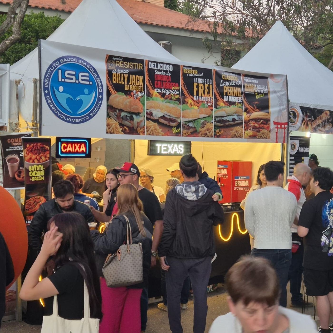 Fila de clientes no caixa da barraca com banners do cardápio