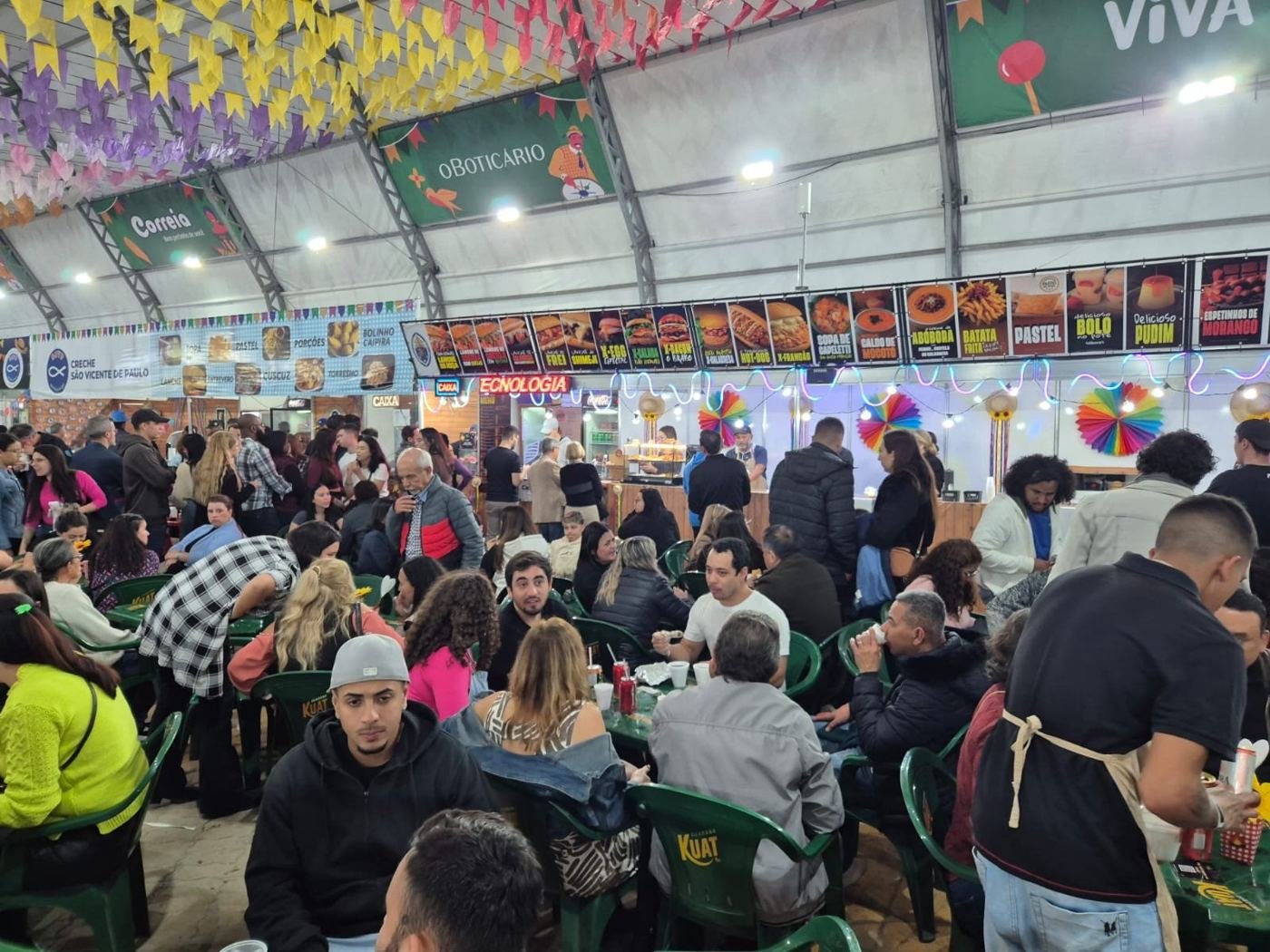 Público lotando as mesas em frente às barracas na festa junina